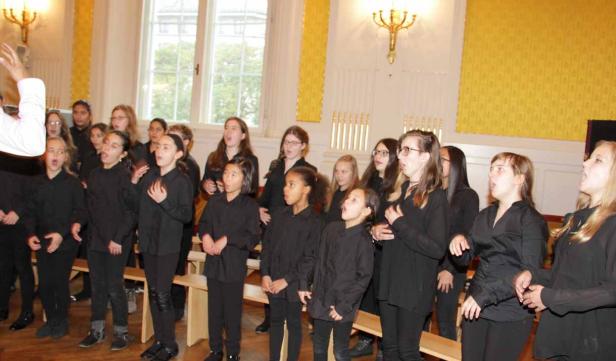 Ein Mädchenchor singt unter der Leitung eines Dirigenten in einem Raum mit gelben Vorhängen.