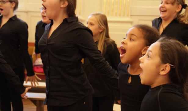Ein Kinderchor probt mit geöffneten Mündern in einem Raum mit Säulen.