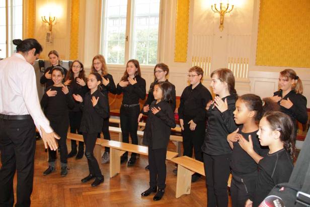 Ein Chor von Jugendlichen und ein Dirigent proben in einem Raum mit gelben Wänden.
