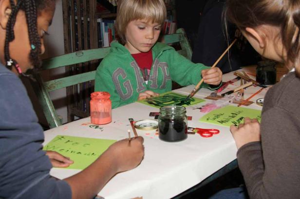 Kinder sitzen an einem Tisch und malen mit Pinseln auf Papier.