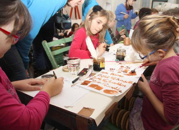 Kinder sitzen an einem Tisch und schreiben mit Feder und Tinte Weihnachtskarten.