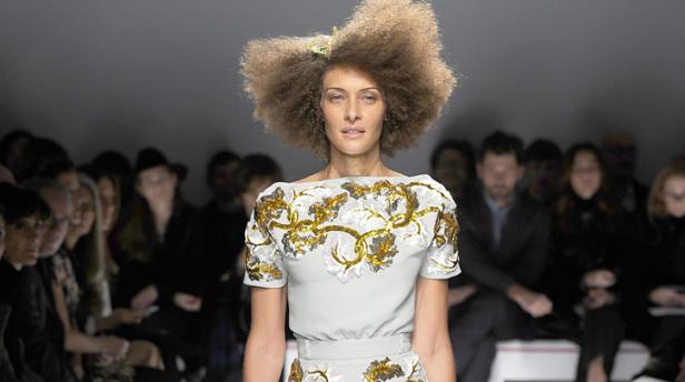 Eine Frau mit Afro-Frisur präsentiert ein hellblaues Kleid mit floralen Applikationen auf einem Laufsteg.