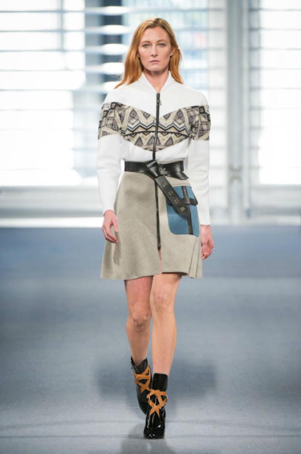 Eine Frau auf dem Laufsteg trägt eine Jacke mit Muster, einen Rock mit Gürtel und schwarze Stiefel.
