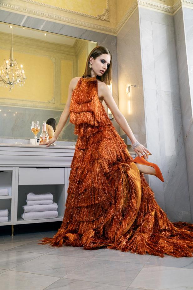 Eine Frau posiert in einem orangefarbenen Fransenkleid und passenden Schuhen in einem Badezimmer.