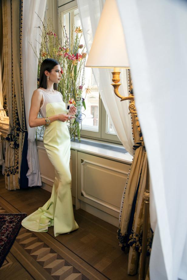 Eine Frau in einem hellgrünen Kleid steht an einem Fenster und hält ein kleines Geschenk.
