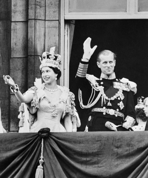 Ein Schwarzweißbild aus London im Jahr 1953: Die Queen steht prunkvoll angezogen nach ihrer Krönung mit ihrem Ehemann auf einem Balkon und wink