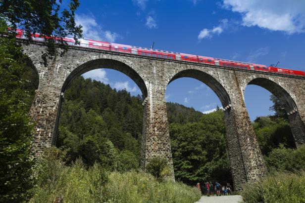 Ein roter Zug fährt über ein steinernes Viadukt, unter dem Wanderer stehen.