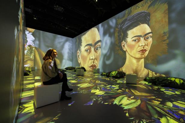 Eine Frau sitzt in einer immersiven Ausstellung mit Projektionen von Frida Kahlo.