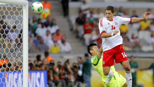 Ein Fußballspieler springt hoch, um einen Ball vor einem Torwart abzuwehren.