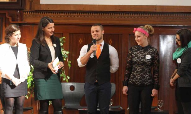 Eine Gruppe von Personen steht auf einer Bühne, ein Mann spricht ins Mikrofon.