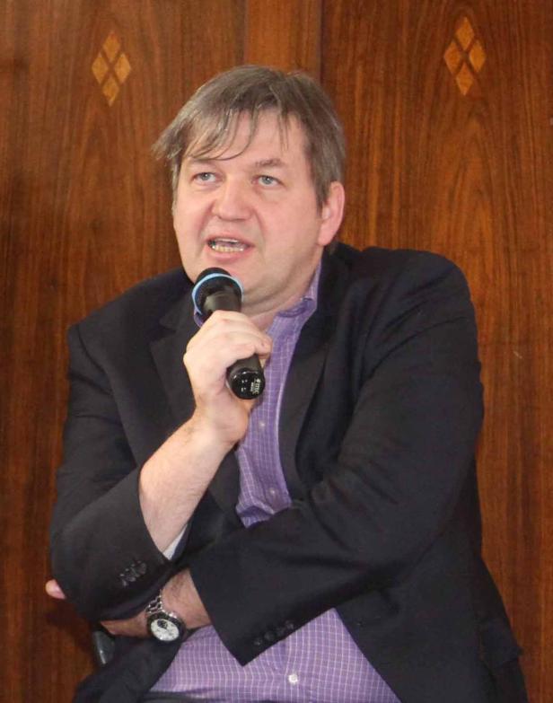 Ein Mann mit Mikrofon spricht vor einer hölzernen Wand.