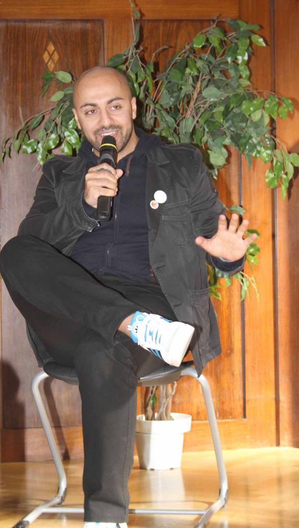 Ein Mann mit Mikrofon sitzt vor einer Holzwand und einer Pflanze.