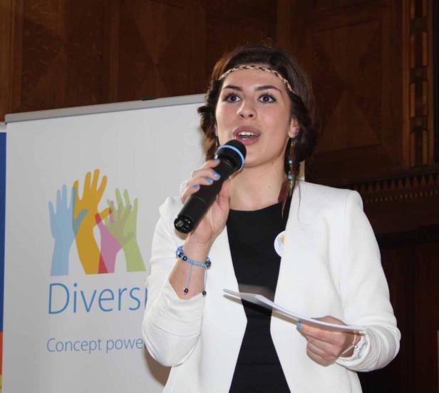 Eine Frau spricht bei einer Veranstaltung ins Mikrofon; im Hintergrund ein Banner mit dem Wort „Diversi“.