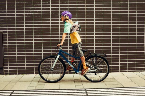 Ein Kind mit lila Helm fährt ein blaues Woom-Fahrrad vor einer Backsteinmauer.