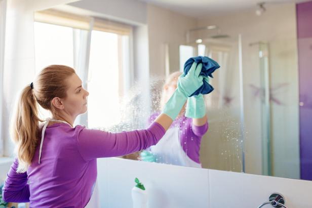 Eine Frau putzt mit einem blauen Lappen und Handschuhen einen Spiegel im Badezimmer.