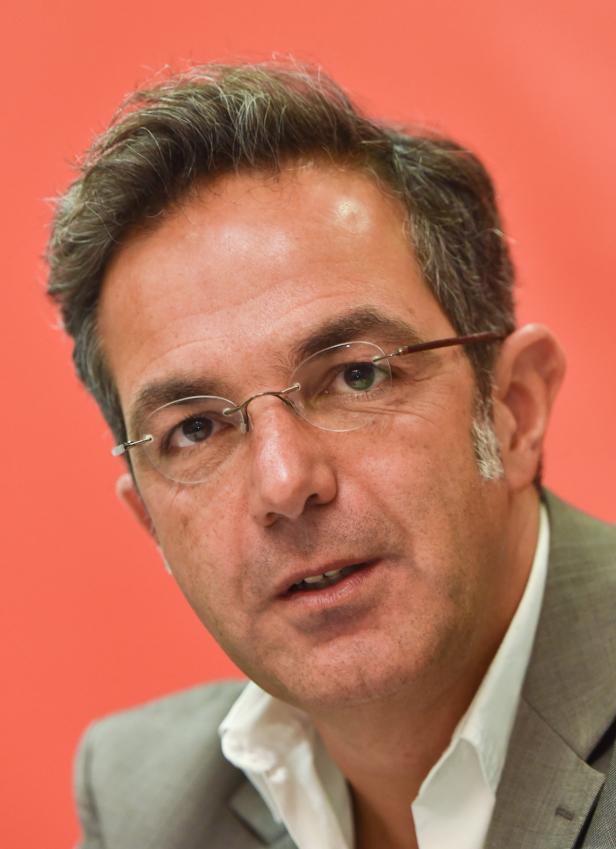 Ein Mann mit Brille und Anzug vor einem roten Hintergrund.