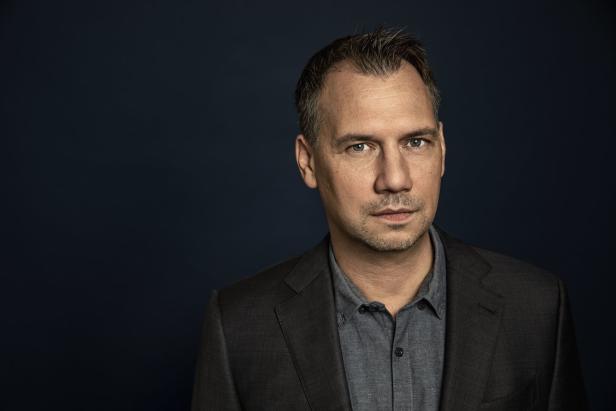 Ein Mann mit Anzug und grauem Hemd vor einem dunklen Hintergrund.