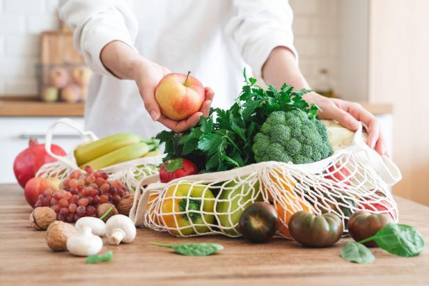 Eine Person hält einen Apfel über eine Netztasche voller Gemüse und Obst.