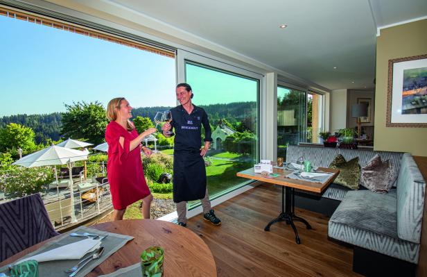 Eine Frau und ein Kellner stoßen in einem Restaurant mit Blick auf eine grüne Landschaft mit Weingläsern an.