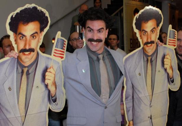 Sacha Baron Cohen, als Borat verkleidet, posiert mit Pappaufstellern seiner Figur, die amerikanische Flaggen halten.
