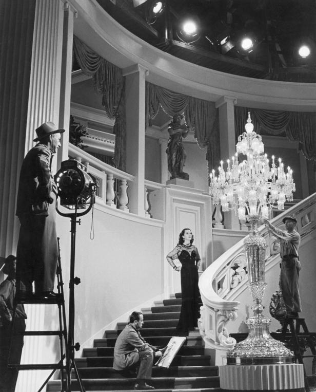 Hedy Lamarr posiert auf einer Treppe während Dreharbeiten, umgeben von Filmcrew und Beleuchtung.
