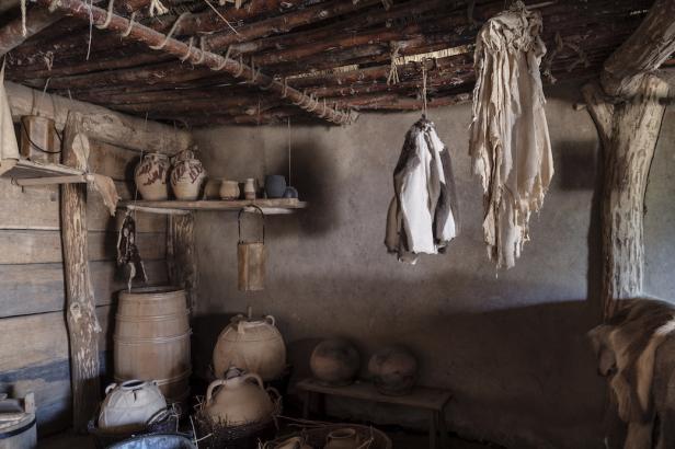 Innenansicht eines historischen Hauses mit Töpferwaren, Fässern und Tierfellen.