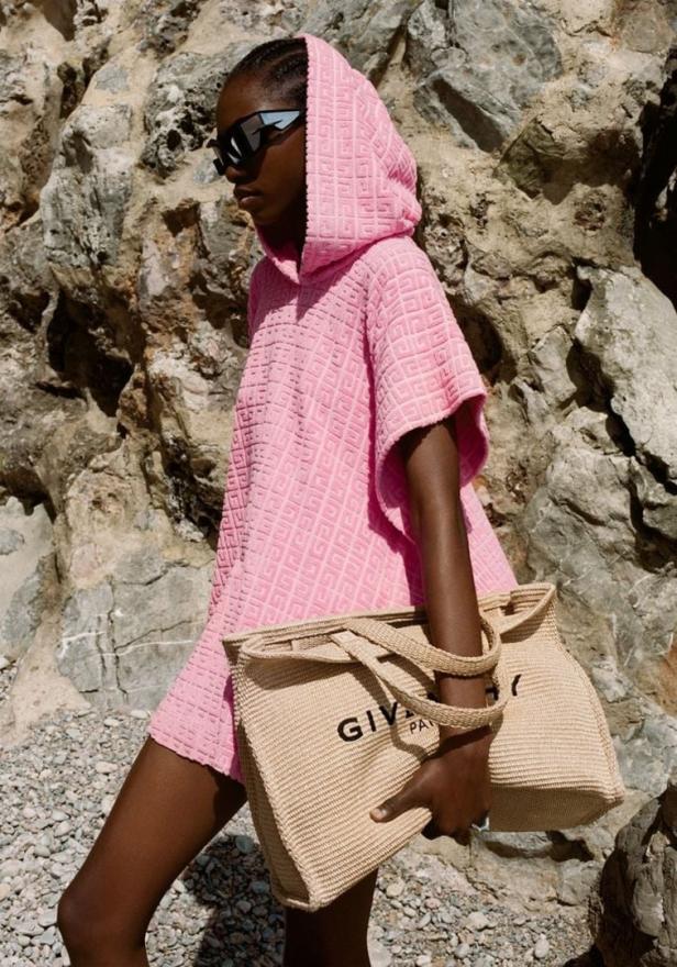 Eine Frau mit Sonnenbrille und einem rosa Givenchy-Strandponcho mit passender Tasche vor einem Felsen.