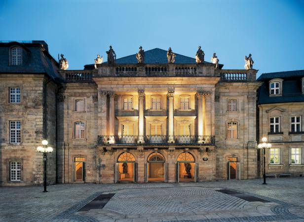Das Neue Schloss Bayreuth mit beleuchteten Fenstern und Statuen auf dem Dach bei Dämmerung.