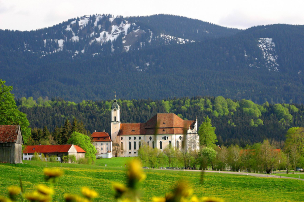 Die Wieskirche vor einer bewaldeten Hügellandschaft mit schneebedeckten Gipfeln.