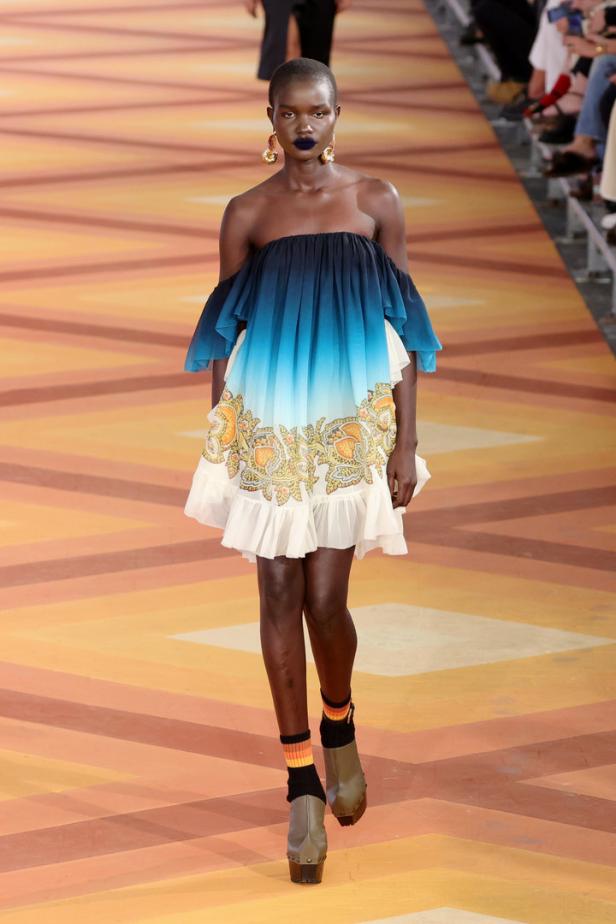 Eine Frau mit dunklem Teint präsentiert ein blau-weißes Kleid mit Blumenmuster auf einem Laufsteg.
