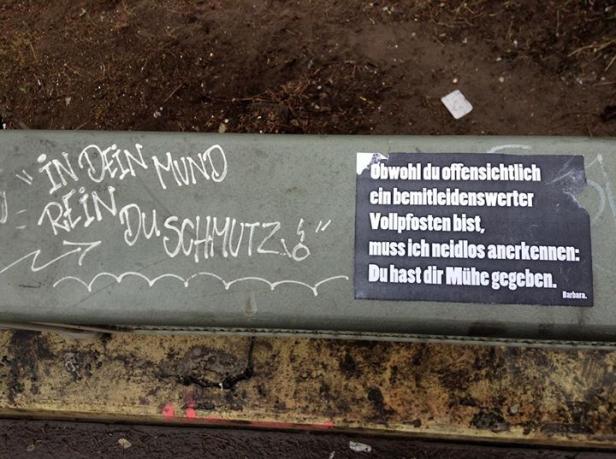 Auf einer Bank befinden sich Graffiti und ein Aufkleber mit dem Text „Obwohl du offensichtlich ein bemitleidenswerter Vollpfosten bist…“.
