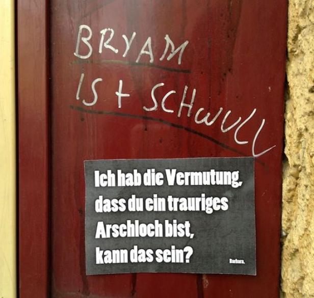 Auf einer roten Tür befinden sich Graffiti und ein Aufkleber mit der Aufschrift „Ich hab die Vermutung, dass du ein trauriges Arschloch bist, kann das sein?“.