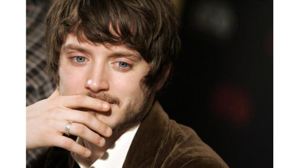 Elijah Wood mit Bart und einem Ring am Finger hält seine Hand vor den Mund.