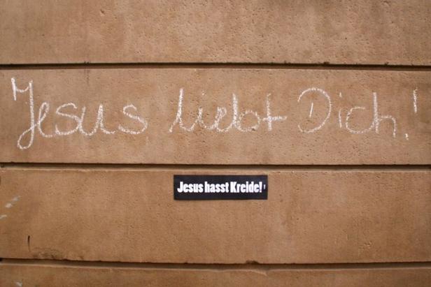 Auf einer Wand steht mit Kreide geschrieben „Jesus liebt Dich!“, darunter ein Aufkleber mit der Aufschrift „Jesus hasst Kreide!“.