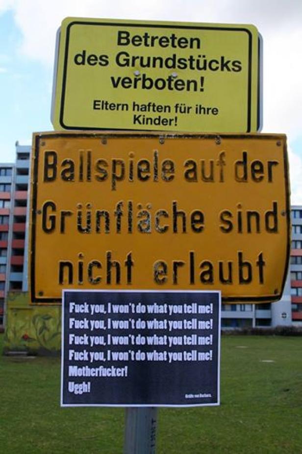 Ein Schild verbietet das Betreten des Grundstücks und Ballspiele auf der Grünfläche; darunter ein Protestplakat.