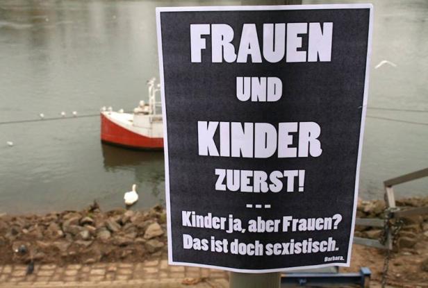 Ein Schild am Flussufer fordert „Frauen und Kinder zuerst!“, kritisiert aber auch Sexismus.
