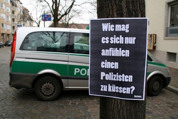 Ein an einem Baum befestigtes Plakat mit der Aufschrift „Wie mag es sich nur anfühlen, einen Polizisten zu küssen?“, im Hintergrund ein Polizeiwagen.