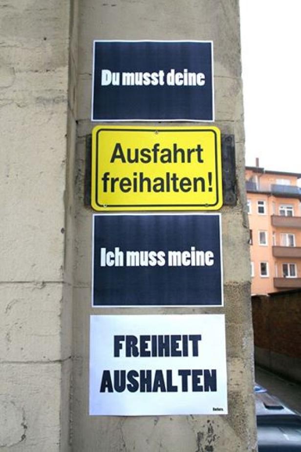 Auf einem Pfeiler sind Schilder mit den Aufschriften „Du musst deine“, „Ausfahrt freihalten!“, „Ich muss meine“ und „Freiheit aushalten“ angebracht.