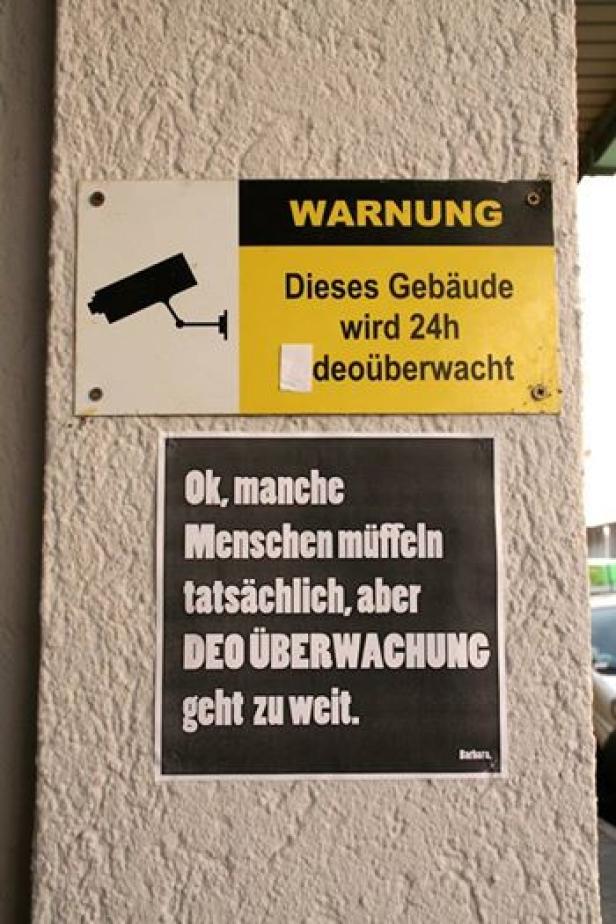 Ein Schild warnt, dass dieses Gebäude 24 Stunden videoüberwacht wird, darunter hängt ein satirischer Kommentar zur „DEO ÜBERWACHUNG“.