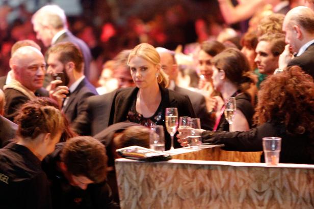 Charlize Theron sitzt an einem Tisch bei einer Veranstaltung, umgeben von anderen Gästen.