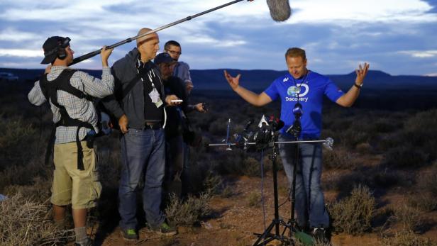 Ein Mann mit blauem Discovery-Shirt gestikuliert vor Mikrofonen, umgeben von einem Filmteam in einer Wüstenlandschaft.