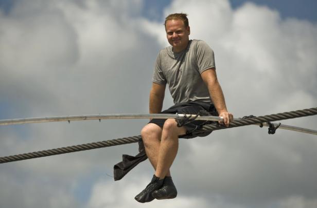 Ein Mann sitzt auf einem Seil vor einem bewölkten Himmel.