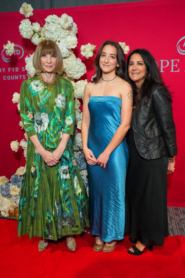 Anna Wintour posiert mit zwei Frauen auf einem roten Teppich vor einem Blumenarrangement.