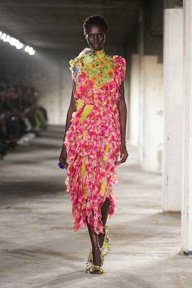 Ein Model präsentiert ein asymmetrisches, mehrlagiges Kleid mit Blumenmuster auf einem Laufsteg.