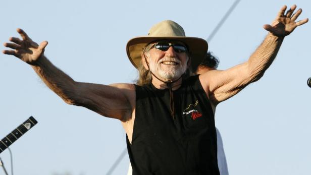 Willie Nelson mit Hut und Sonnenbrille steht mit erhobenen Armen auf der Bühne.