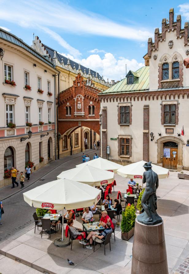 Blick auf einen Platz in Krakau mit Cafés, Sonnenschirmen und einer Statue.