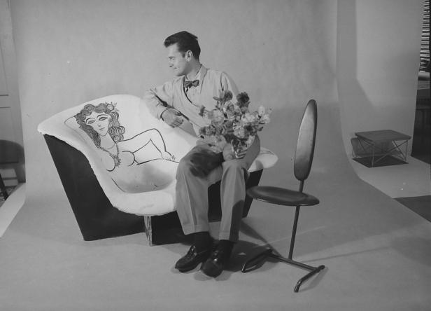 Ein Mann mit Fliege sitzt auf einem Sofa mit einer aufgemalten Frau und hält einen Blumenstrauß.