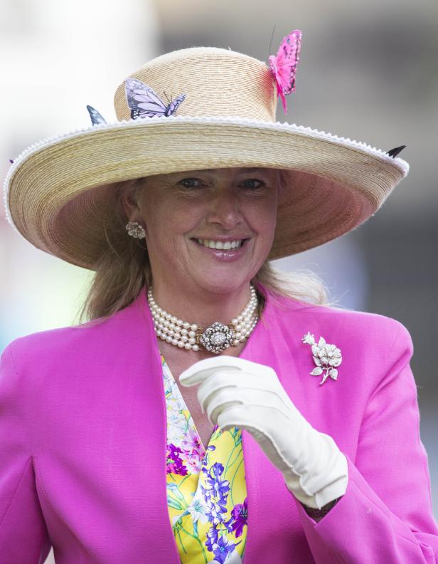 Eine Frau mit Hut, verziert mit Schmetterlingen, und einem pinkfarbenen Blazer lächelt.