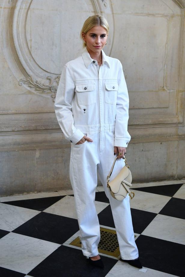 Eine Frau in einem weißen Overall mit einer Dior-Tasche vor einem verzierten Hintergrund.