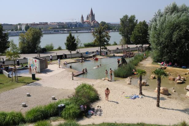 Ein Badesee mit Sandstrand und spielenden Menschen in Wien, im Hintergrund die Kirche St. Franziskus.